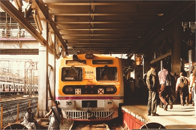 [ai] A yellow train at a busy train station platform with passengers boarding and disembarking. Other trains are visible in the background, and the station is covered with a metal roof.